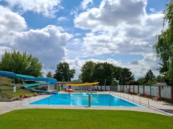 Freibad mit Schwimmbecken, Rutsche und Liegewiese unter blauem Himmel.